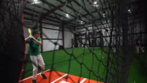 Indoor turf arena with sports equipment and a batter getting ready to hit a ball surrounded by netting