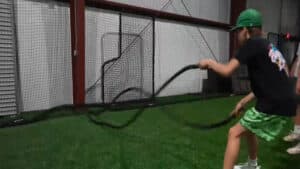 Indoor turf arena with sports equipment and netting surrounding a person lifting heavy ropes for exercise