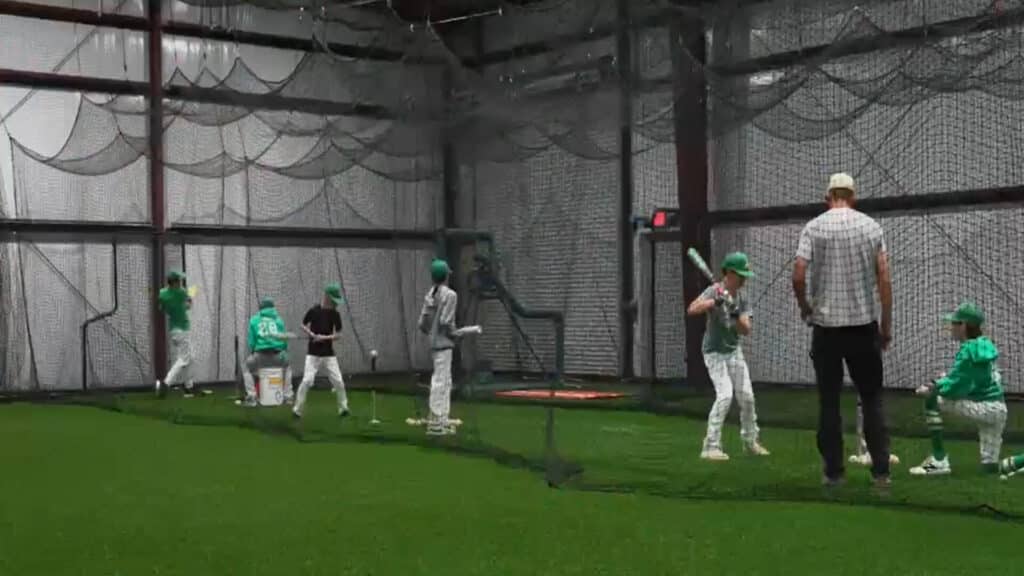 Indoor turf arena with sports equipment and netting with batters and coaches practicing hitting baseballs